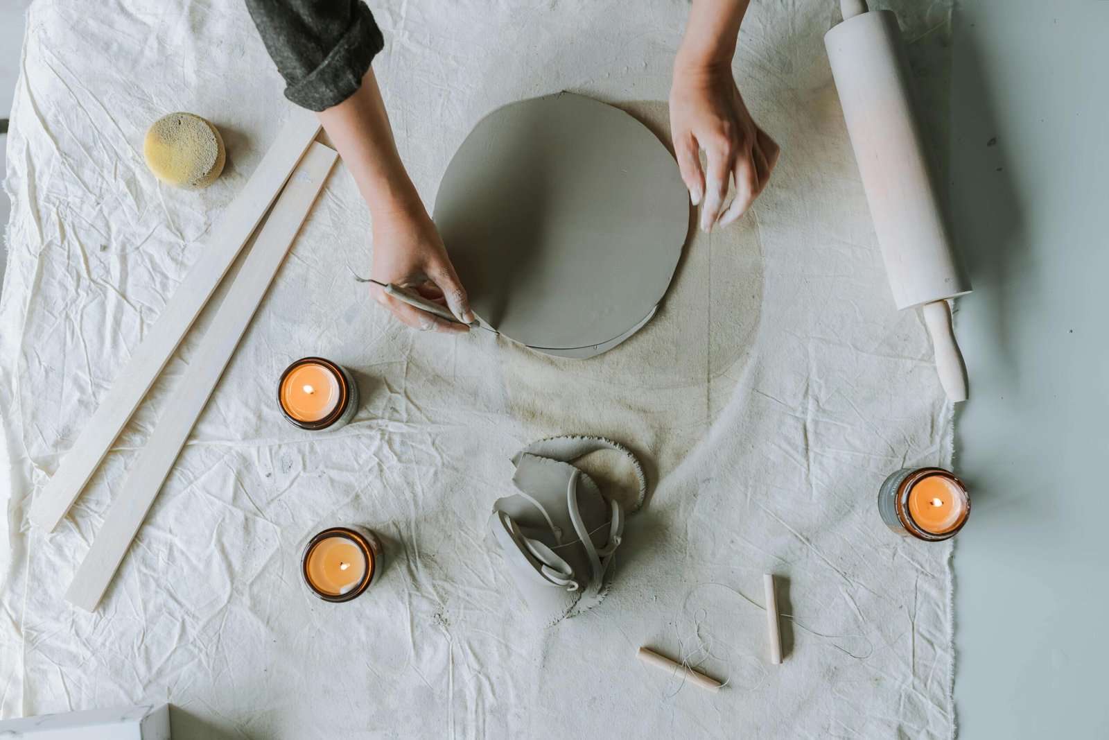 Top view of hands crafting clay with candles and tools on table, creating a peaceful mood.